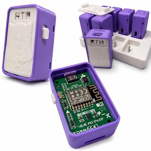Puede incluir: Un dispositivo electrónico rectangular de color morado con una placa frontal blanca que presenta un personaje de anime en relieve y el texto TOMAMCAT. El dispositivo contiene una placa de circuito verde con un chip ESP-12F, un puerto USB y el texto WEAR AND ENJOY.