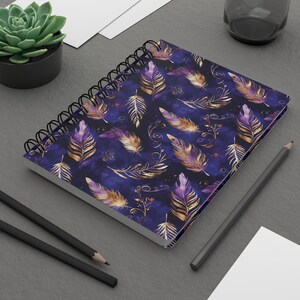 May include: Spiral-bound notebook featuring a dark blue cover adorned with a pattern of gold and purple feathers. The notebook is open, revealing blank pages. Two black pencils and a small potted succulent are also visible.