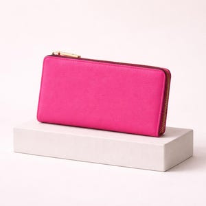 Könnte beinhalten: Eine leuchtend pinkfarbene rechteckige Geldbörse mit einem goldfarbenen Reißverschluss und Zipper. Die Geldbörse wird auf einem weißen rechteckigen Block vor einem weißen Hintergrund präsentiert. Der Reißverschluss verläuft entlang drei Seiten der Geldbörse.