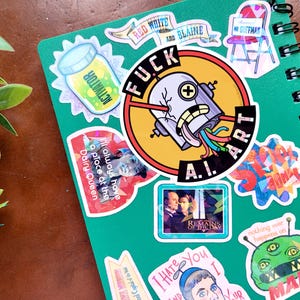 Puede incluir: Un cuaderno verde en espiral cubierto de pegatinas coloridas. Las pegatinas incluyen el texto "FUCK ART A.I.", "Red White and Blaine" y un extraterrestre de dibujos animados. Otras pegatinas presentan varios dise&ntilde;os y texto.