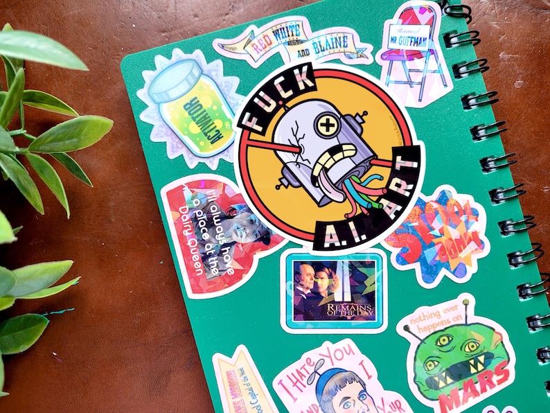 Puede incluir: Un cuaderno verde en espiral cubierto de pegatinas coloridas. Las pegatinas incluyen el texto "FUCK ART A.I.", "Red White and Blaine" y un extraterrestre de dibujos animados. Otras pegatinas presentan varios dise&ntilde;os y texto.