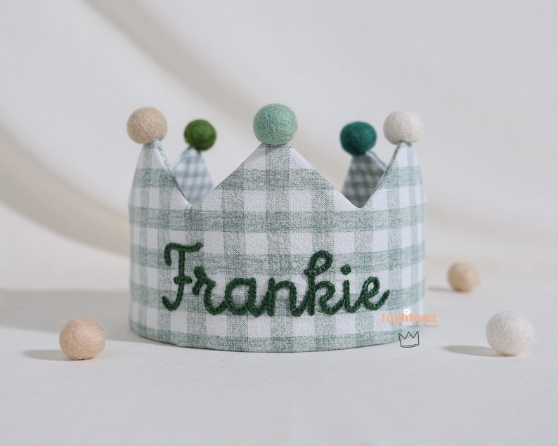 K&ouml;nnte beinhalten: Eine Stoffkrone mit gr&uuml;n-wei&szlig;em Vichy-Muster, bestickt mit dem Namen "Frankie" in Gr&uuml;n. Die Krone ist mit kleinen, bunten Pompons verziert. Kleine beige und wei&szlig;e Filzkugeln sind um die Krone herum verstreut.