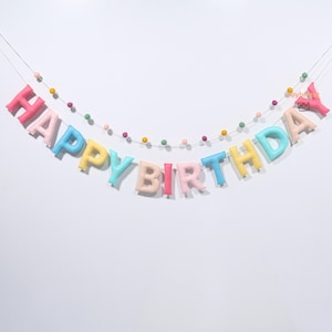 Può includere: Un festone colorato "Happy Birthday" con lettere dai colori pastello e una fila di piccole perline rotonde. Le lettere sono rosa, blu, giallo e altre tonalità tenui, creando un aspetto festoso e allegro. Perfetto per una festa di compleanno.
