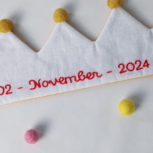 May include: A white fabric crown featuring yellow felt pom-poms on each point and red embroidered text that reads 02 - November - 2024. The crown includes a white elastic band and is accented by loose pink and yellow felt pom-poms.