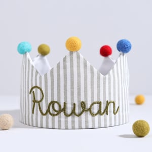 Peut inclure: Une couronne en tissu avec un motif à rayures verticales grises et blanches. Le prénom Rowan est brodé en écriture cursive verte sur le devant. Les pointes supérieures sont ornées de petits pompons en feutrine colorés bleu, jaune, rouge et bleu sarcelle.