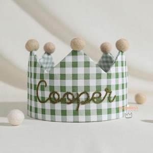 May include: A fabric crown with a green and white gingham pattern. The word "Cooper" is embroidered in green thread across the front. The crown is topped with beige felt balls.