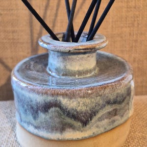 May include: A ceramic essential oil diffuser with a light brown base and a cylindrical body in shades of blue, grey, and brown. Black reeds are inserted into the top opening, creating a decorative and functional home fragrance item.