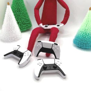May include: A red fabric doll with white gloves holding a miniature white and black video game controller. Two additional identical miniature white and black video game controllers are positioned in front of the doll, surrounded by small cone-shaped trees.