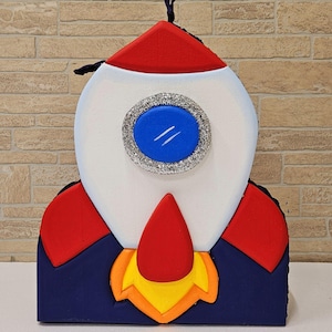May include: A colorful rocket ship decoration with a red nose cone, red fins, and a red top. The body is white with a blue and silver porthole. The base is navy blue with orange and yellow flames.
