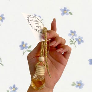 May include: A small, clear glass bottle filled with a golden liquid, suspended by a gold cord and wooden bead. The bottle has a wooden cap. A white tag with the text "Coffee Shop" is attached. The background is white with blue floral patterns.