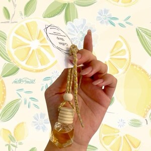 Peut inclure: Petite bouteille d'aromathérapie en verre transparent remplie d'un liquide brun clair, suspendue par un cordon doré et une perle en bois. L'étiquette indique "Coffee Shop Aromatherapy". Fond floral et citron.