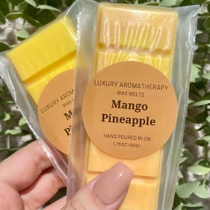 May include: Two wax melts in clear plastic packaging. The wax melts are yellow and have a label that says "Luxury Aromatherapy Wax Melts Mango Pineapple Hand Poured in on 1.75oz (50g)"