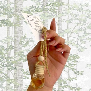 May include: A small, clear, hexagonal glass bottle filled with a light brown liquid, suspended by a gold cord and wooden accents. A white tag with the text "Coffee Shop" hangs from the cord. The background features a light green and white tree pattern.