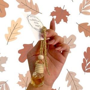 May include: A small, clear glass aromatherapy bottle with a wooden cap and a gold cord. The bottle contains a light brown liquid. A white tag with the text "Coffee Shop" is attached. The background features a pattern of brown and tan leaves.