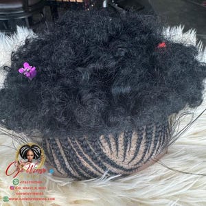 Puede incluir: Una peluca negra rizada con una base trenzada, adornada con una pequeña flor morada y un acento rojo. La peluca se muestra sobre una superficie blanca y esponjosa. El logotipo de Ojo Weave Wigs es visible.