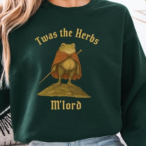 May include: Dark green sweatshirt featuring the text "Twas the Herbs M'lord" and a cartoon frog in a cape, holding a staff. The frog is on a mound. The sweatshirt has a crew neck design.