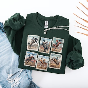 May include: A dark green sweatshirt featuring six vintage-style postage stamp designs with images of cowboys riding bucking broncos. A silver necklace with a circular pendant is draped across the sweatshirt. The sweatshirt is paired with ripped blue jeans.