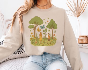 Könnte beinhalten: Beiges Sweatshirt mit einer skurrilen Illustration einer Parkszene mit zwei Figuren, Bäumen, Schmetterlingen und Blumen. Das Design ist in einem weichen, Vintage-Stil gehalten, ideal für Freizeitkleidung.