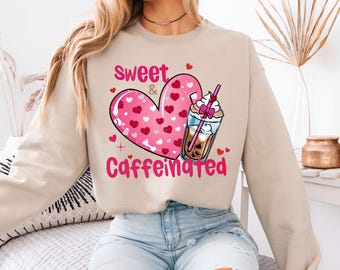 Könnte beinhalten: Beiges Sweatshirt mit einem rosa Herzdesign, das mit kleineren Herzen gefüllt ist. Die Worte "Sweet & Caffeinated" sind in Rosa gedruckt. Eine Grafik eines Kaffeegetränks mit Schlagsahne ist ebenfalls abgebildet.