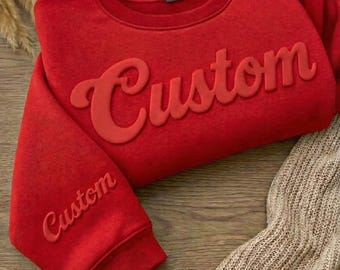 May include: A red sweatshirt with the word "Custom" in a raised, matching color font across the chest and on the left sleeve. The sweatshirt is folded on a wooden surface, with a beige knitted fabric visible in the background.