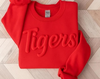 Puede incluir: Una sudadera roja brillante con la palabra "Tigers" en relieve, en cursiva. La sudadera tiene cuello redondo y mangas largas con puños acanalados. La etiqueta de la marca Gildan es visible en el interior del cuello.