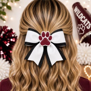 May include: A hair bow featuring a white center ribbon with black glitter edges, accented by a maroon paw print emblem in the middle. The bow is constructed with layered fabric loops and long, pointed tails, creating a structured accessory.