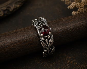 Puede incluir: Un anillo de plata con una gran gema roja oscura facetada en el centro. El anillo está adornado con intrincados detalles de hojas y vides. El anillo se muestra sobre una superficie de madera oscura, mostrando su elegante diseño.