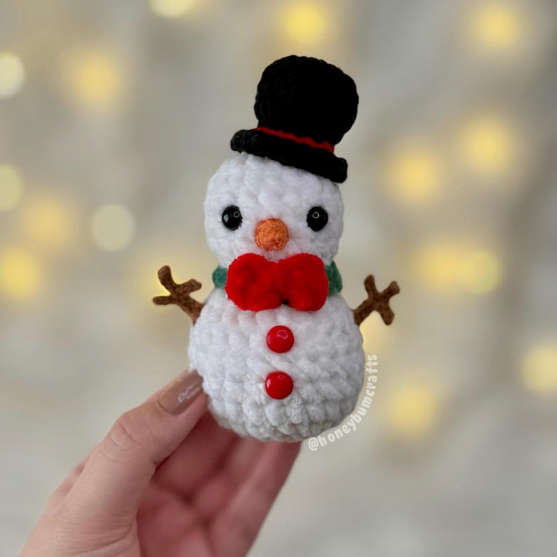 May include: A crocheted snowman with a black top hat, a red bow tie, and two red buttons. The snowman has a carrot nose and twig arms. The snowman is white with black eyes and a green scarf.
