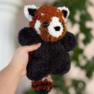 May include: A handmade plush red panda toy. The toy features a fluffy black body, a reddish-brown head and back, and white accents on the face and ears. The red panda has a small black nose and black eyes. The toy is held in a person's hand.