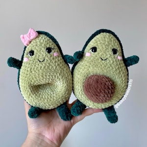 Puede incluir: Dos juguetes de aguacate de peluche, uno con un lazo rosa, el otro con un hueso marrón. Ambos tienen caras sonrientes, mejillas rosadas y detalles en verde oscuro. Los juguetes se sostienen en una mano, mostrando su apariencia suave y texturizada.