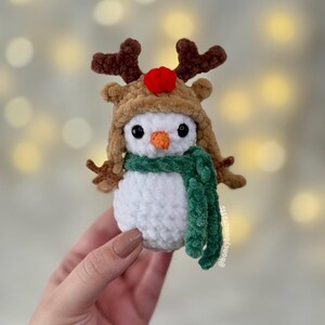 May include: A handmade crochet snowman wearing a reindeer hat with brown antlers and a red pom-pom. The snowman has a green scarf and an orange carrot nose. The snowman is a festive holiday decoration.