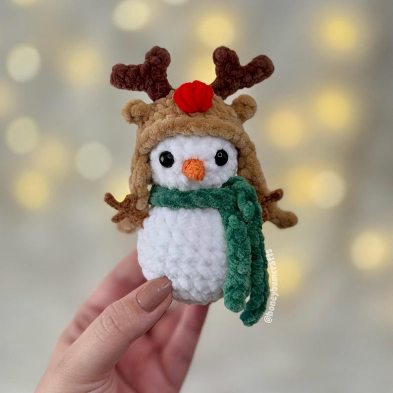 May include: A handmade crochet snowman wearing a reindeer hat with brown antlers and a red pom-pom. The snowman has a green scarf and an orange carrot nose. The snowman is a festive holiday decoration.