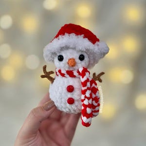 May include: A handmade crochet snowman with a red and white Santa hat and scarf. The snowman has black eyes, an orange nose, and brown twig arms. Two red buttons adorn the snowman's body. The background is blurred with bokeh lights.