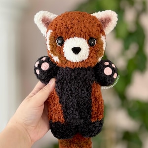 Könnte beinhalten: Ein handgefertigtes gehäkeltes Rotpanda-Plüschtier. Das Plüschtier hat einen braunen und schwarzen Körper, weiße Gesichtsmerkmale und rosa Pfoten. Die Augen sind groß und schwarz. Die Ohren sind weiß mit rosa Akzenten. Das Plüschtier wird in einer Hand gehalten.