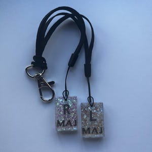 May include: A black lanyard with a silver clasp and two rectangular glittery charms. The charms have the letters "R" and "L" with the word "MAJ" below each letter. The charms are attached to the lanyard with black cords.