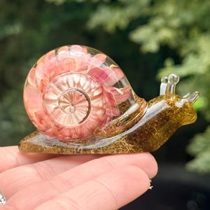 May include: A clear resin snail figurine featuring a spiral shell filled with pink flower petals. The body of the snail is tinted a translucent amber color and contains embedded golden moss-like fibers throughout its structure.