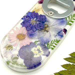 Puede incluir: Un abridor de botellas de resina transparente con un abridor de acero inoxidable. El abridor está incrustado con flores secas en tonos morados, rosas y blancos. El abridor tiene un pequeño agujero para colgar.