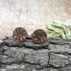May include: A pair of copper cufflinks with a crossed arrow design. The cufflinks are round and have a rustic finish.