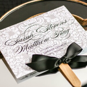 May include: A white and gray wedding program fan with black calligraphy text that reads "The Wedding of Sarah Matthew and Steven Gray, October 15, 2015, St. Dennis Church, Freeport, Illinois". A black satin ribbon is tied around the fan handle.