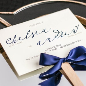 May include: A white wedding programme with navy blue calligraphy for the wedding of Chelsea Hope Hughes and Andrew Michael Maicus. The programme is tied with a navy blue satin ribbon and a wooden stick.