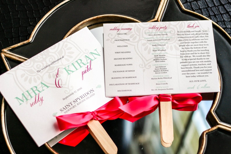 May include: Wedding ceremony program fans with a pink ribbon and wooden stick. The program includes the wedding ceremony schedule, wedding party names, and a thank you message to guests. The front of the fan features the names "Mira Kiran Reddy" and the date "September 12, 2016".
