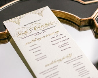 May include: A wedding ceremony program with gold lettering on a cream background. The program lists the order of events for the wedding ceremony of Ruth and Christopher, which will take place on October 5, 2015 at Saint Dennis Church in Lockport, Illinois.