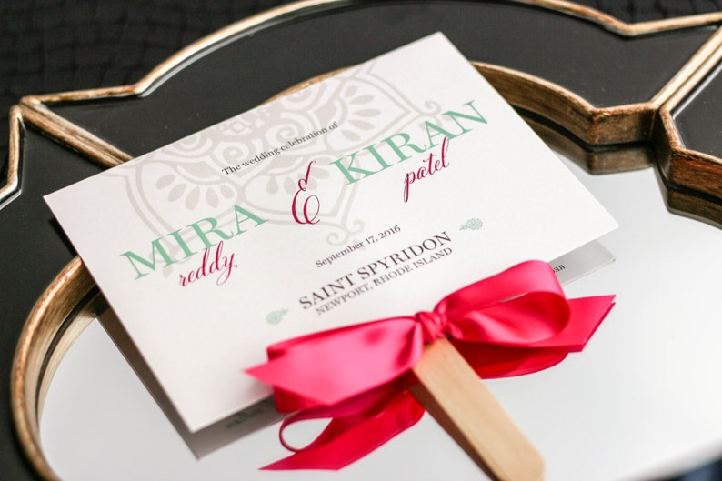 May include: A white wedding program with a teal and gray patterned design. The program is titled "The wedding celebration of Mira Reddy & Kiran Patel" and includes the date "September 17, 2016" and the location "Saint Spyridon, Newport, Rhode Island". The program is attached to a wooden stick with a bright pink bow.