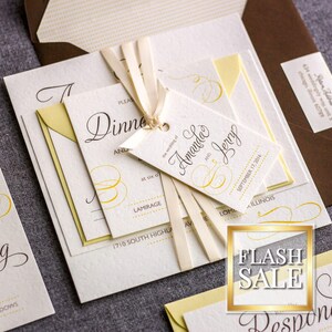 Op de afbeelding: Elegante trouwuitnodigingssuite met crèmekleurige kaarten, gele accenten en een bruine envelop. De uitnodiging heeft kalligrafie en een label met de namen "Amanda en Jerry". Een "Flash Sale"-bord is zichtbaar.