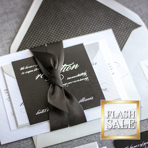 May include: Elegant wedding invitation suite featuring a black card with white calligraphy, tied with a black satin ribbon. The suite includes white and gray envelopes with patterned linings, and additional cards. A gold-framed "FLASH SALE" graphic is visible.