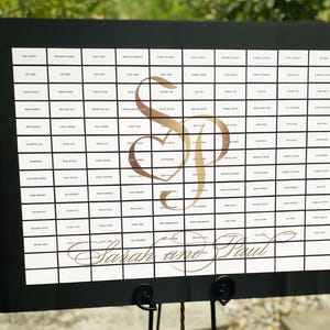 Könnte beinhalten: Ein schwarz gerahmtes Schild mit einem Raster aus kleinen Kästchen, die jeweils Text enthalten. In der Mitte befindet sich ein goldenes Monogramm mit den Buchstaben "S" und "P", die mit einem Herzen verschlungen sind. Die Worte "Sarah and Paul" sind kursiv geschrieben.