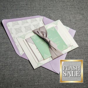 May include: A set of wedding invitations with a lavender envelope, white cardstock, and a green inner card. The cards are tied together with a gray satin ribbon.