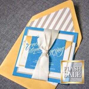 May include: Wedding invitation suite featuring a blue and white invitation card with elegant script, tied with a white ribbon. The suite includes a gold envelope and a striped liner. A gold-framed "FLASH SALE" sticker is visible.