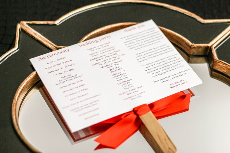 May include: A white wedding ceremony program with red text, folded into a fan shape with a red ribbon and wooden handle. The program lists the order of the ceremony and the wedding party.