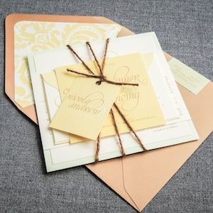 Puede incluir: Un conjunto de invitaciones de boda con un sobre amarillo y marrón, una tarjeta crema con una etiqueta amarilla que dice "Nicole y Andrew", y una tarjeta amarilla con el texto "Boda de verano".